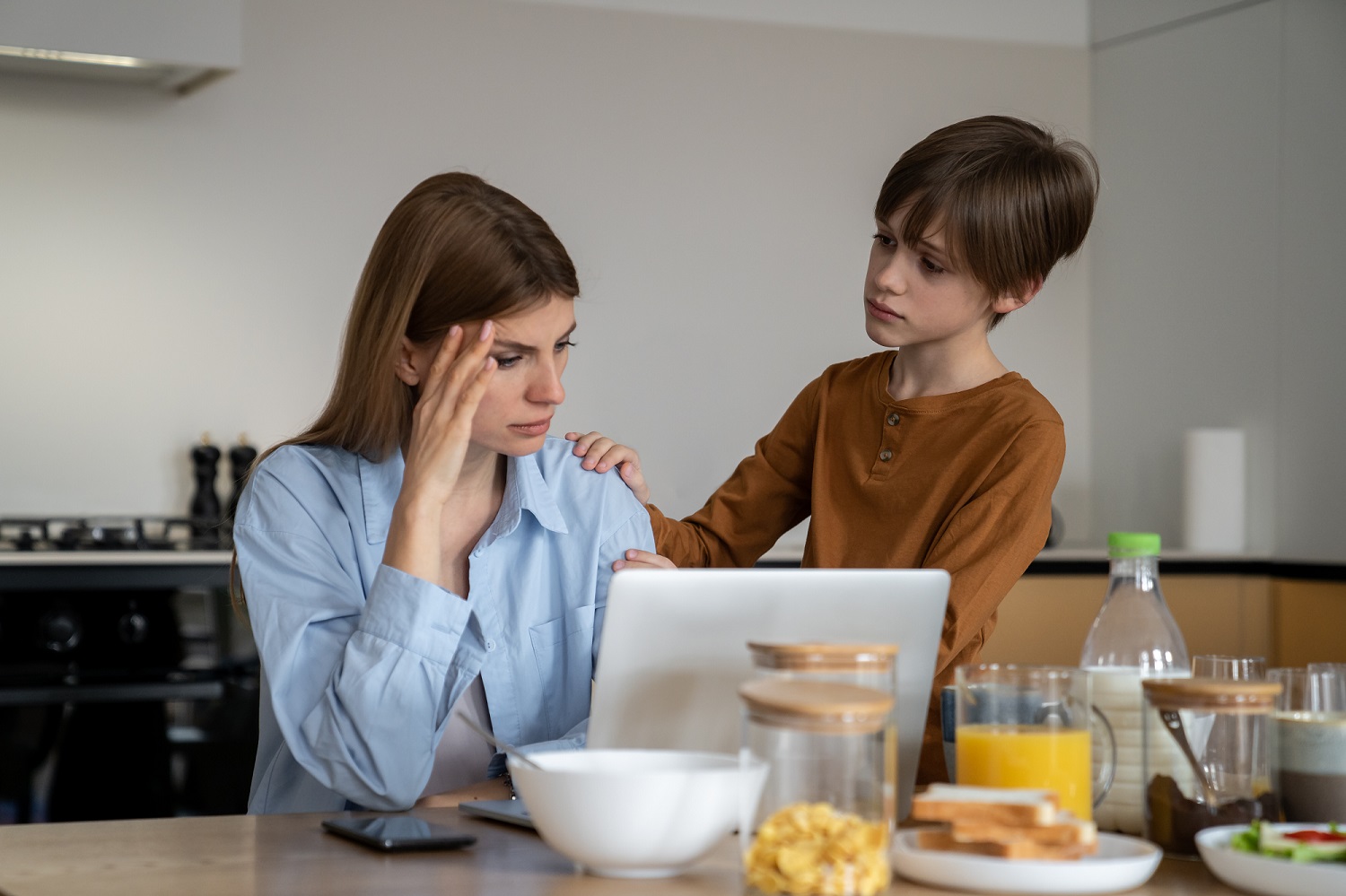 Vrouw zit bezorgd achter haar laptop met haar zoon rechts van haar.